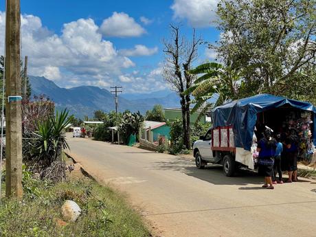 A rural street scene in a small village in Chiapas, with a vendor truck parked on the side of the road. Local women dressed in traditional clothing stand by the truck, while the background features mountains and greenery under a partly cloudy sky.