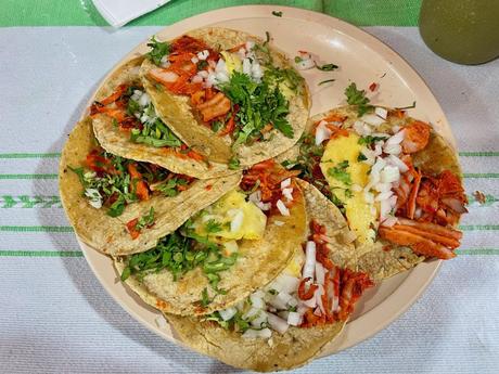 A plate of tacos al pastor with pineapple, onion, and cilantro on soft corn tortillas, served on a simple plate over a striped tablecloth.