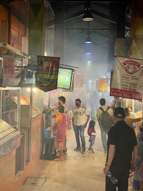 A bustling indoor food market in Oaxaca City filled with smoke from grilled meats, with vendors preparing food and customers walking through narrow aisles beneath hanging banners and signs.