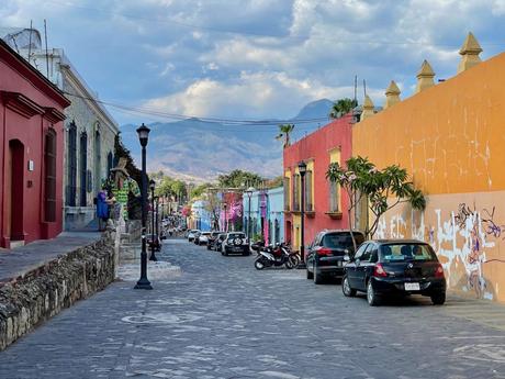 A quiet street in Oaxaca lined with colorful colonial-style buildings, with mountains visible in the distance under a partly cloudy sky.