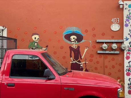 A mural of two skeletons in traditional Mexican attire painted on a bright orange wall, with a red pickup truck parked in front.