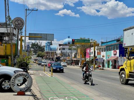 A bustling street in Oaxaca with cars and motorcycles, blue domed buildings in the background, and a sign pointing towards Oaxaca city center.