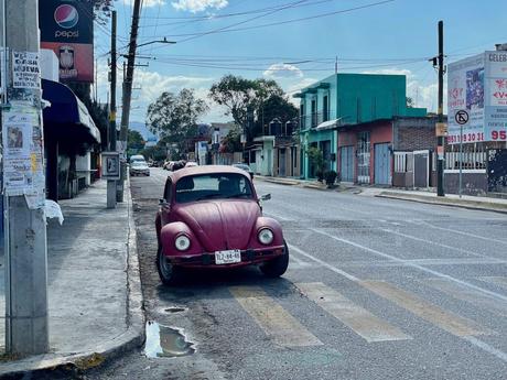 A red Volkswagen Beetle is parked along a street lined with vibrant buildings, while a Pepsi sign and various storefronts can be seen in the background.