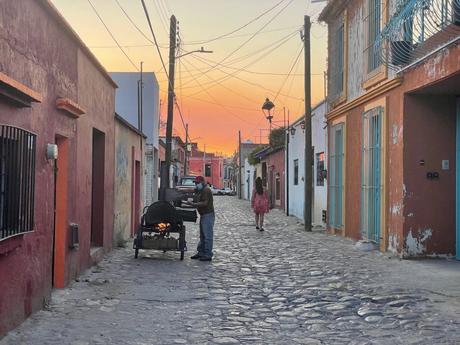 A narrow cobblestone street at sunset, with warm orange and pink hues in the sky. A man stands by a grill with a fire, cooking, while a woman in a dress walks down the street. The houses are painted in vibrant colors like red, orange, and teal. Power lines stretch overhead, adding to the rustic charm of the scene.