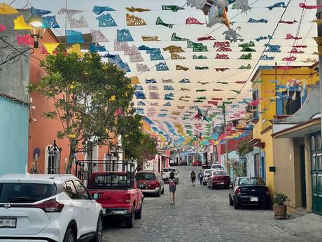 A narrow street decorated with colorful papel picado banners overhead, lined with brightly painted buildings and cars parked on both sides.