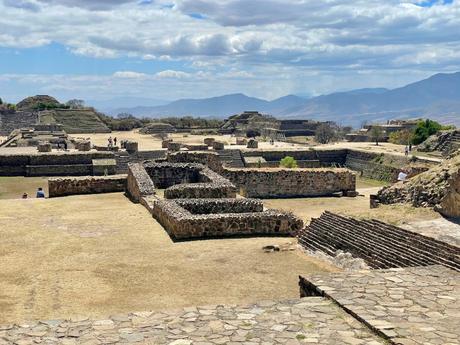 Stone ruins at Monte Albán, with various walls and structures surrounded by mountainous terrain, tourists exploring the area.