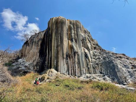 A towering petrified waterfall formation at Hierve el Agua, rising dramatically from the mountainside, with a few people resting at the base.