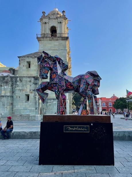 A colorful sculpture of two horses made from geometric stained glass stands on display in front of a historic stone church under a clear blue sky in Oaxaca.