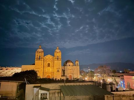 The historic Santo Domingo church in Oaxaca, lit up at night against a cloudy sky, with its intricate stone façade glowing warmly.