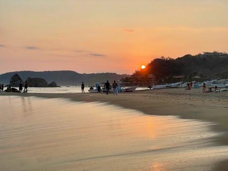 A sunset scene on sandy San Agustinillo Beach in Oaxaca, Mexico, with silhouettes of people and boats along the shore, and hills in the background.