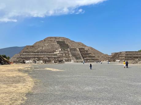 A view of the Pyramid of the Sun at Teotihuacan, with visitors walking on the wide open space in front of the ancient structure under a bright blue sky.