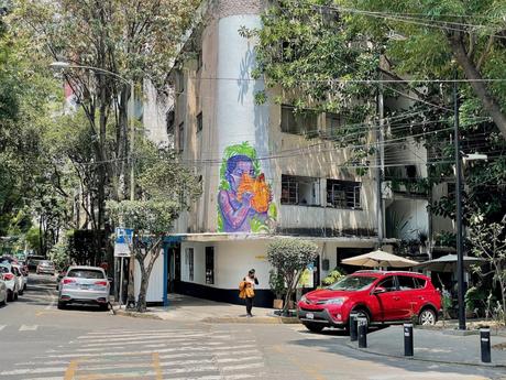 A tall building with a vibrant mural of a person blowing into an orange flower, set in the leafy Condesa neighborhood with parked cars and a pedestrian crossing.