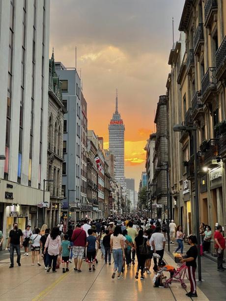 A bustling street scene at sunset with people walking, framed by tall historic buildings, and the Torre Latinoamericana illuminated in the background.