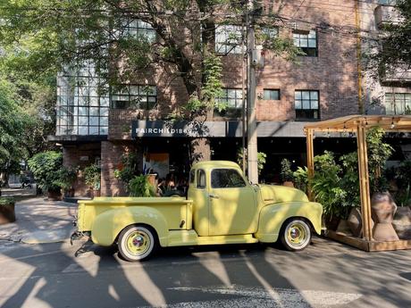 A vintage yellow truck parked in front of a modern café building in a leafy street, casting shadows from nearby trees in Mexico City.