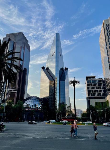 Skyscrapers in Mexico City, including the striking triangular Torre Reforma, rising against a blue sky with clouds, surrounded by palm trees.