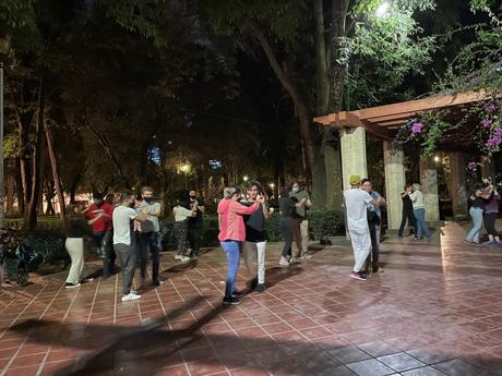 A group of people, some wearing masks, dancing salsa together in a park at night, under trees with soft lighting.