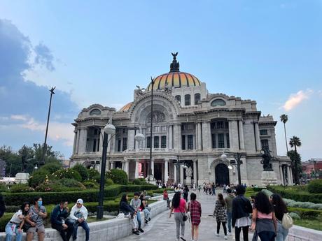 The front of Palacio de Bellas Artes with its majestic dome and marble facade, as people sit and walk in the plaza surrounding it.