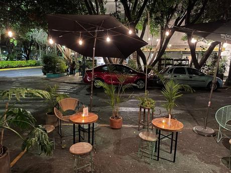 A small outdoor cafe setting at night with round tables and chairs under an umbrella, surrounded by greenery and lit with hanging light bulbs.