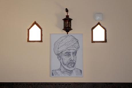 A framed sketch of Sultan Qaboos in traditional Omani headgear hangs on a wall between two small arched windows, with a traditional lantern suspended above.