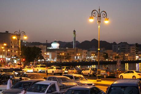 A cityscape of Muscat, Oman, at dusk, with cars filling the streets, and a mosque with a domed roof and minaret lit up against the backdrop of mountains.