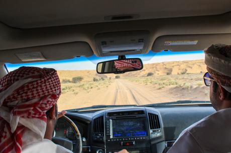 Inside a car with two Omani men driving through a sandy desert, seen through the windshield, with the barren landscape stretching ahead.