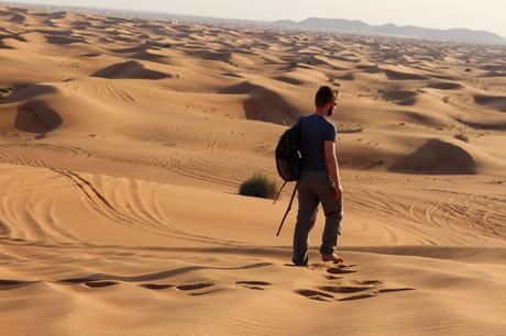 Alex Tiffany wearing a backpack walks barefoot across golden sand dunes, leaving footprints behind, with rolling dunes stretching into the horizon.