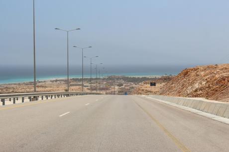 A wide-open highway leading down to the turquoise waters of the Arabian Sea, with desert landscapes on either side of the road.