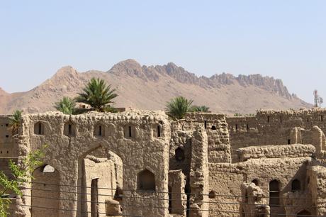 Ancient mud-brick ruins with small windows, surrounded by palm trees, against the backdrop of jagged mountains under a bright sky.