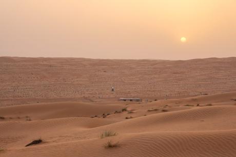 A distant view of the sun setting over the vast Omani desert, casting a soft orange glow over the sand dunes, with a small structure visible on the horizon.