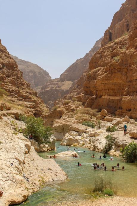 A clear, refreshing pool of water in Wadi Shab, Oman, surrounded by rugged, steep brown cliffs, with people swimming and enjoying the natural setting.