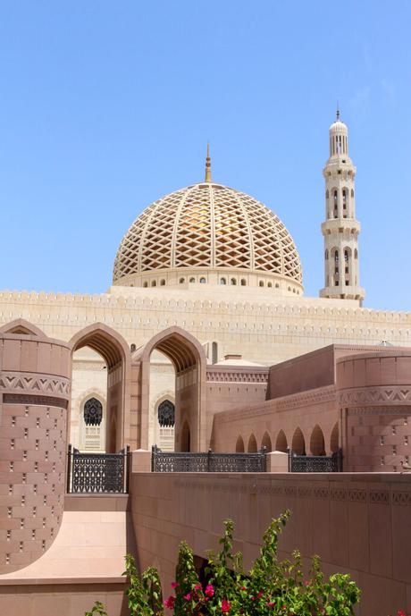 A panoramic view of the Sultan Qaboos Grand Mosque, showcasing its grand dome, minaret, and beautifully landscaped surroundings, set against a clear blue sky.