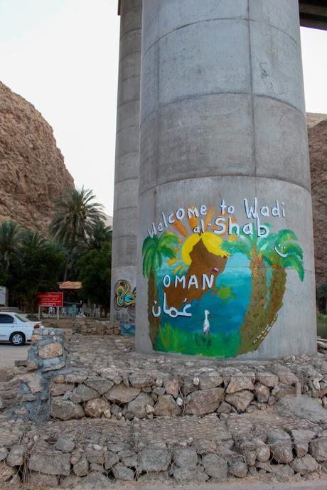 A painted mural on a concrete pillar at Wadi al-Shab, Oman, welcoming visitors with colorful imagery of a sun, palm trees, and a heron.