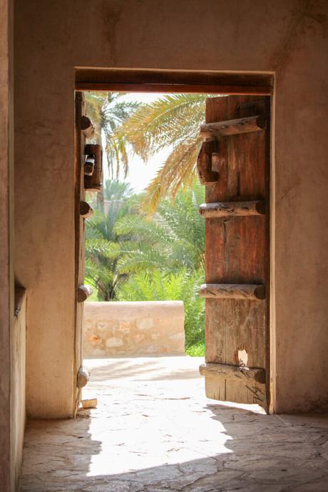 The view through an open wooden door framed by thick walls, looking out at bright sunlight illuminating palm trees and stone structures.