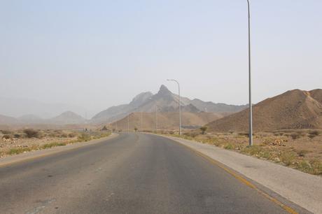 A winding road through Oman’s desert landscape, with distant mountains under a hazy sky and streetlights lining the side of the road.