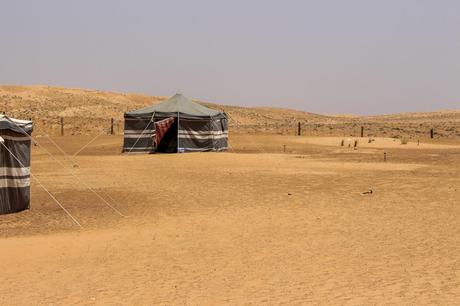 Two traditional Bedouin tents in the Omani desert, made of striped fabric, standing on the sandy ground with the arid, rolling dunes in the background.