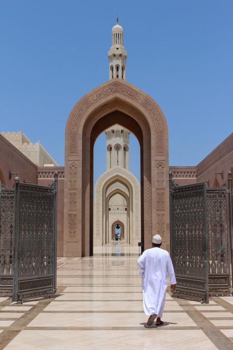 A man dressed in traditional Omani clothing walks through a series of intricately designed archways at a mosque, with a tall minaret visible in the background.