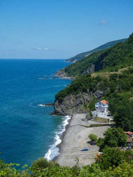 A scenic coastal view of the Black Sea in Turkey, with a rocky shoreline, a white building perched on a cliff, and vibrant blue water stretching to the horizon.