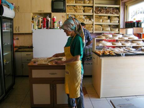 An elderly woman in a headscarf and apron stands in a small shop or bakery, preparing food on a countertop surrounded by shelves filled with packaged goods and bread.