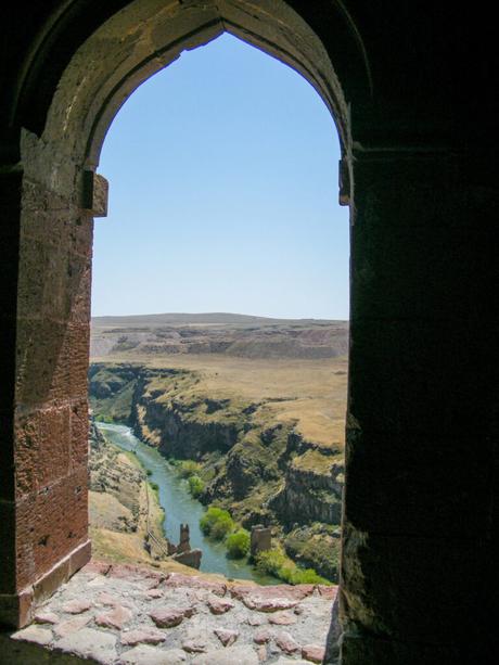 A picturesque view through an arched stone window of a flowing river cutting through a rugged canyon, with a bright blue sky above.