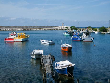 Several small fishing boats floating in a peaceful harbor with a stone pier and lighthouse in the background, under a bright blue sky.