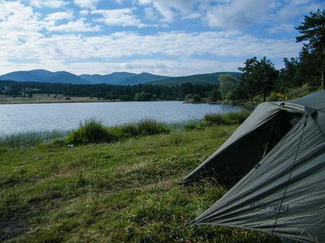 A peaceful campsite by a lake surrounded by grassy fields and hills in the background, with a green tent set up in the foreground under a blue sky with fluffy clouds.