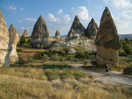 Unique rock formations resembling fairy chimneys in Cappadocia, Turkey, with a lone hiker walking through the arid landscape dotted with trees.