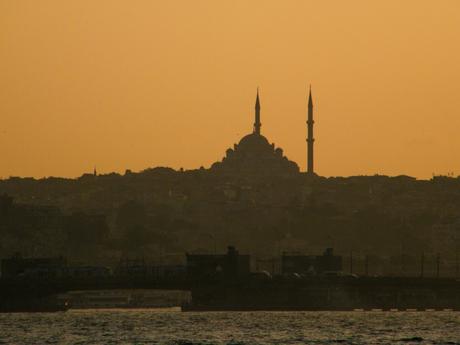 The silhouette of a mosque with two minarets against an orange sunset sky in Istanbul, Turkey, creating a dramatic contrast with the darkened cityscape below.