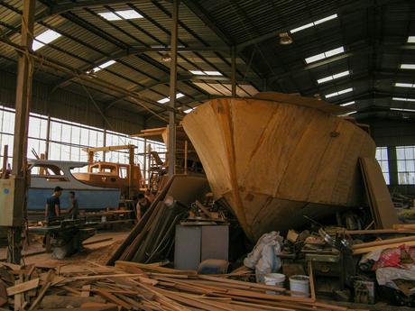 An interior view of a boat workshop, showing a large wooden boat under construction with workers and tools scattered around.