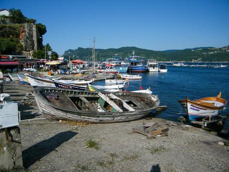 A bustling harbor with various fishing boats and small vessels docked along a pier, with nearby cafes and stalls under bright blue skies.