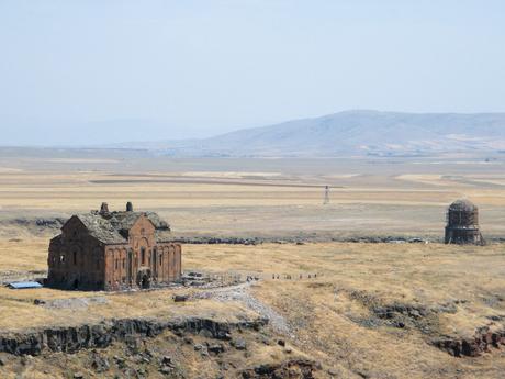 A closer look at the Ani Cathedral and a smaller domed building, set against a backdrop of rolling hills and open plains.