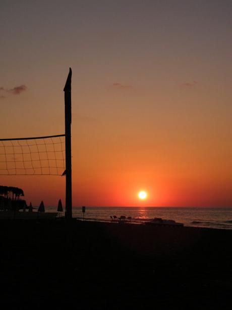 A beach volleyball net silhouetted against a stunning orange sunset over the sea, with a few beach chairs and umbrellas visible in the distance.