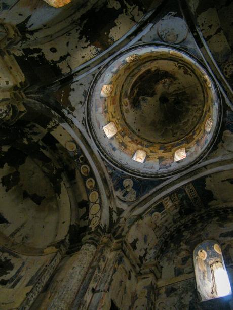 The interior of a deteriorating old church, with faded frescoes on the walls and ceilings, and natural light filtering through small windows.