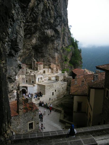 A view of the historic Sumela Monastery, nestled against a steep cliff face, with visitors exploring the ancient stone buildings and courtyards.