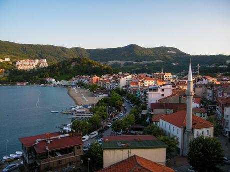 A scenic view of a coastal town in Turkey with red-roofed houses, a mosque with twin minarets, and a green hillside in the background, overlooking a calm bay.